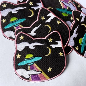 May include: A set of black and white embroidered iron-on patches featuring a cat silhouette with a UFO, stars, and a crescent moon. The patches have pink stitching around the edges.