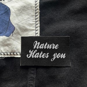 May include: A black fabric patch with white text that reads "Nature Hates You". The patch is sewn onto a black fabric background. A white fabric square with a cartoon image of a girl is sewn onto the fabric background above the patch.