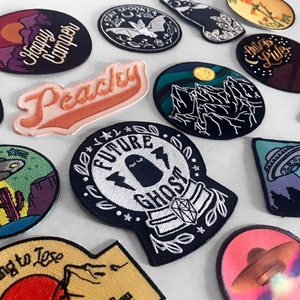 May include: A collection of embroidered iron-on patches featuring various designs, including a ghost in a crystal ball, a peach, a mountain range, a UFO, and a happy camper. The patches are all black and white with colorful accents.