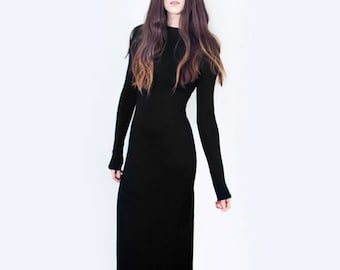 May include: A floor-length, solid black dress featuring long sleeves and a high, rounded neckline. The garment is fitted through the torso and falls in a straight, column-like silhouette to the floor, made of a smooth, opaque fabric.