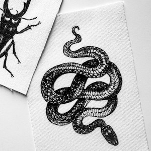 May include: Black and white illustrations of a beetle and a snake on white fabric patches. The beetle is depicted with large mandibles, while the snake is coiled. The artwork has a detailed, vintage style.