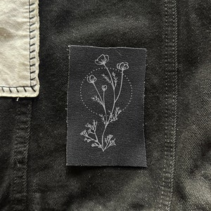 May include: A black fabric patch with a white outline of a flower with five petals and a stem with leaves. The flower is surrounded by a circle with dots.
