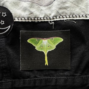 May include: A green luna moth with long, thin wings and a white body is depicted on a black fabric patch. The patch is sewn onto a black denim jacket.
