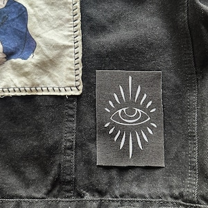 May include: A black denim jacket with a white eye patch sewn on the front. The eye patch has a white outline and a white sunburst design.