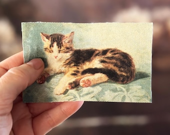 Könnte beinhalten: Ein rechteckiger Aufnäher mit einer detaillierten Illustration eines Tabby-Kätzchens. Das Kätzchen hat braune und schwarze Streifen und weiße Markierungen. Der Hintergrund ist hellblau mit einem floralen Muster.