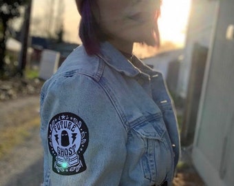May include: A person wearing a blue denim jacket with a black and white patch that says "Future Ghost" on the left sleeve. The person is also wearing a red beanie and glasses.