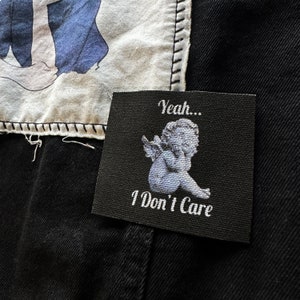 May include: Black patch with white text and a white image of a cherub. The text reads "Yeah... I Don't Care."