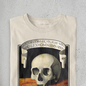 K&ouml;nnte beinhalten: Cremefarbenes T-Shirt mit einer detaillierten Sch&auml;delgrafik und einem Banner mit lateinischem Text. Das Design ist in einem schwarzen Rahmen eingefasst. Der Markenname "ESPI/LANE" ist oben aufgedruckt.