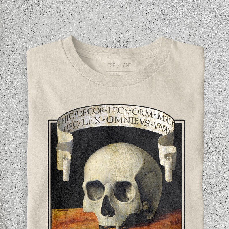 K&ouml;nnte beinhalten: Cremefarbenes T-Shirt mit einer detaillierten Sch&auml;delgrafik und einem Banner mit lateinischem Text. Das Design ist in einem schwarzen Rahmen eingefasst. Der Markenname "ESPI/LANE" ist oben aufgedruckt.