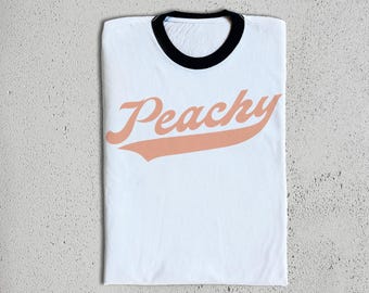 Op de afbeelding: Wit T-shirt met een zwarte ringelhals en een perzikkleurige "Peachy"-afbeelding in een vintage baseballlettertype.