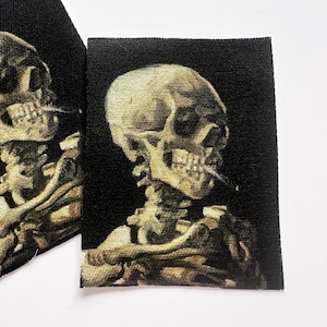 May include: Two fabric patches featuring a skull with a cigarette in its mouth, rendered in shades of beige and brown against a black background. The patches are rectangular and have a slightly textured appearance, suitable for sewing or ironing onto fabric.