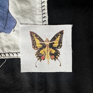May include: A yellow and black butterfly with a human-like figure in the center. The butterfly is on a white fabric background.