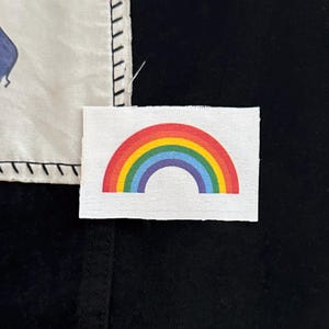 May include: A small, rectangular patch with a rainbow design. The rainbow features bands of red, orange, yellow, green, blue, and purple. The patch is on a white background, with a black background in the lower portion of the image.
