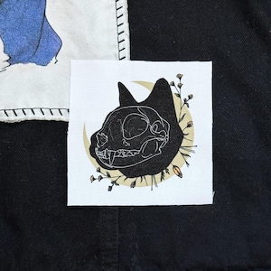 May include: Square fabric patch featuring a black cat skull design with a crescent moon and floral accents. The patch is on a black background, with a white border and stitched edges. The design is detailed and artistic.