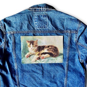 May include: Blue denim jacket with a rectangular patch featuring a vintage illustration of a tabby cat resting on a blue surface. The cat has brown and white fur.