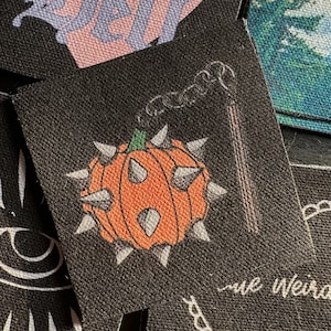 May include: A black patch with a cartoon image of a spiky orange pumpkin with a green stem and a wisp of smoke coming from the top. The text "The Weird" is written in white at the bottom of the patch.