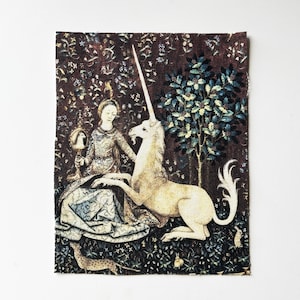 May include: A tapestry featuring a woman and a unicorn in a forest setting. The woman is seated, holding a mirror, with the unicorn resting beside her. The background is a dark brown with floral and tree details, and the unicorn is a creamy white color.