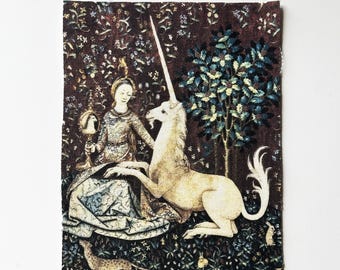 Könnte beinhalten: Ein Wandteppich mit einer Frau und einem Einhorn in einem Wald. Die Frau sitzt und hält einen Spiegel, neben dem Einhorn. Der Hintergrund ist dunkelbraun mit floralen und baumartigen Details. Das Einhorn ist cremeweiß.