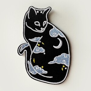 May include: Black embroidered patch of a cat with a crescent moon and clouds. The cat is curled up with its tail wrapped around its body. The clouds are white and the moon is white with a black crescent shape.