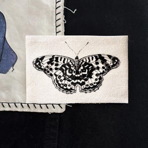 May include: A small, rectangular fabric patch featuring a detailed black and white illustration of a butterfly. The butterfly has intricate patterns on its wings. The patch is set against a background of other fabric pieces.
