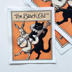 May include: Square fabric patch with the text "The Black Cat" above an illustration of a black cat and a white rabbit playing banjos on an orange background. The patch has a white border.