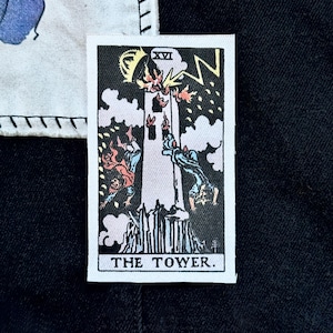 May include: A rectangular patch featuring a tarot card illustration. The card depicts a tower struck by lightning, with figures falling. The text "THE TOWER" is at the bottom, with "XVI" at the top. The color palette includes black, white, red, and gold.
