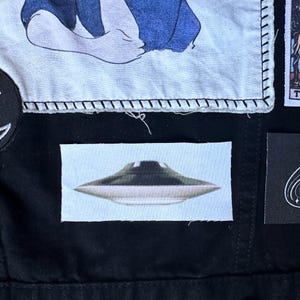 May include: A white patch with a black stitched border featuring a gray and white UFO against a light blue background.