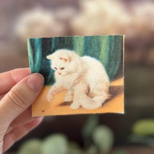 May include: A small, square print featuring a fluffy white kitten with pink accents. The kitten is sitting on a yellow surface with a green and blue background. The print is held by a hand.
