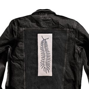 May include: Black denim jacket with a rectangular patch featuring a detailed black centipede design. The jacket has a classic collar, button closure, and pockets, offering a casual and edgy aesthetic.