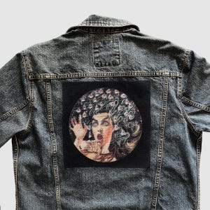 May include: A denim jacket with a black circular patch featuring a portrait of a woman with a surprised expression and floral hair. The jacket is a faded blue color with visible stitching and a button-down front.
