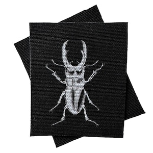 May include: A black fabric patch with a white embroidered stag beetle. The beetle has large mandibles and long legs.