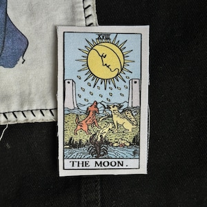 May include: A rectangular fabric patch featuring a tarot card illustration titled THE MOON. The design includes a yellow sun with a face, two towers, a red dog, a yellow wolf, and a lobster emerging from blue water, all on a light background.