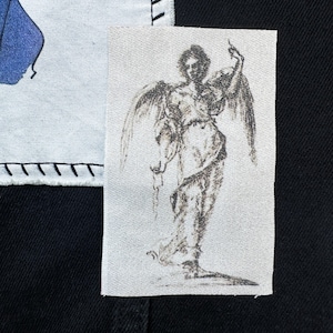 May include: A black and white drawing of an angel with wings outstretched, printed on a white fabric patch.