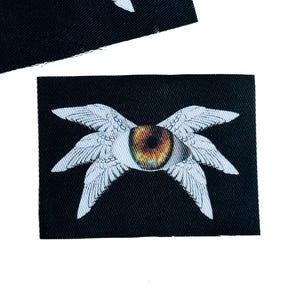 May include: A rectangular black fabric patch featuring a central graphic of a single human-like eye with a brown and gold iris, surrounded by four white feathered wings extending outward in a symmetrical pattern against the dark background.