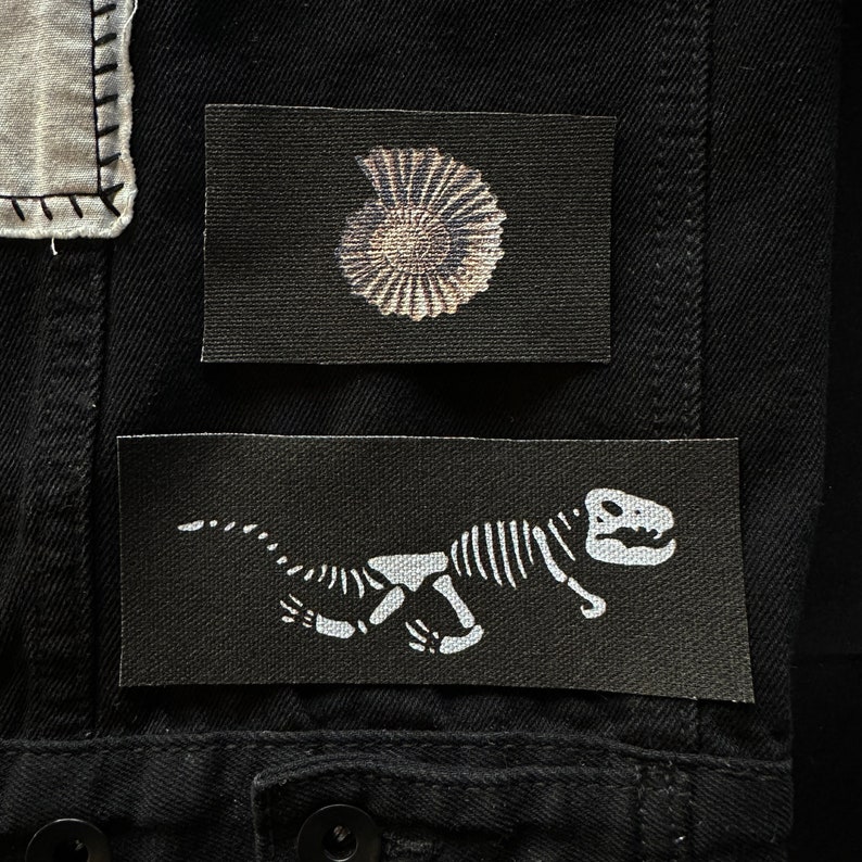 May include: Two black fabric patches with white designs. The first patch features a white ammonite fossil. The second patch features a white dinosaur skeleton.