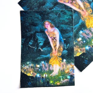 May include: A rectangular fabric patch featuring a printed illustration of a person with long hair wearing a yellow dress, standing in a dark forest setting surrounded by small glowing lights and figures near the ground.