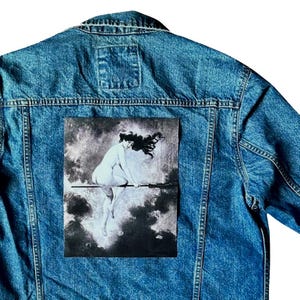 May include: A medium-wash denim jacket with a black and white image of a nude woman riding a broomstick. The artwork is centered on the back of the jacket, with the denim's seams and pockets visible.