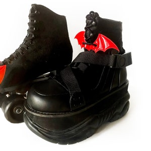 K&ouml;nnte beinhalten: Schwarze Plattform-Rollschuhe mit einem roten Fledermausfl&uuml;gel-Anh&auml;nger, der an der Kn&ouml;chelriemen befestigt ist. Die Schuhe haben eine dicke Plattformsohle und ein schwarzes Schn&uuml;rdesign.