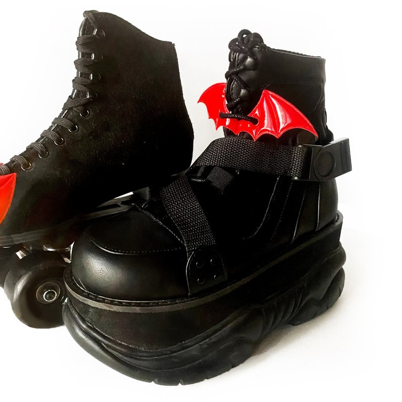 K&ouml;nnte beinhalten: Schwarze Plattform-Rollschuhe mit einem roten Fledermausfl&uuml;gel-Anh&auml;nger, der an der Kn&ouml;chelriemen befestigt ist. Die Schuhe haben eine dicke Plattformsohle und ein schwarzes Schn&uuml;rdesign.