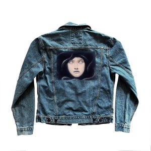 May include: A blue denim jacket with a rectangular patch on the back featuring a portrait of a woman with pale skin and white eyes. The jacket has a classic design with button closures and pockets.