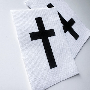 May include: Two white fabric patches with black crosses. The crosses are different sizes and are centered on the patches.