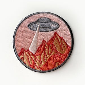 May include: Embroidered patch featuring a gray UFO hovering over a red mountain range against a pink sky. The UFO has a white beam of light shining down on the mountains.