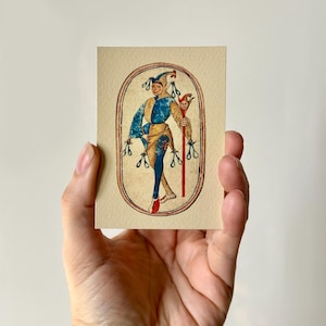 May include: A vintage-style illustration of a jester in blue, yellow, and red attire, holding a staff with a small face. The artwork is framed within an oval border on a cream-colored card, held in a hand.
