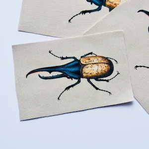 May include: A rectangular fabric patch featuring a detailed illustration of a rhinoceros beetle. The beetle has a dark blue, elongated horn and a textured, tan-colored wing casing, set against a plain, light beige, woven fabric background.
