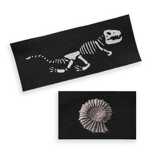 May include: Two black fabric patches with white designs. The first patch features a dinosaur skeleton. The second patch features a fossilized ammonite.