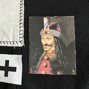May include: A close-up of a portrait of Vlad the Impaler, a historical figure, on a black fabric background. The portrait is a printed image on a fabric patch. The portrait shows Vlad wearing a red and gold crown and a red and gold robe.