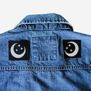 May include: A denim jacket with two black patches featuring a white crescent moon and three stars design. The patches are square and placed on the back of the jacket, near the collar.