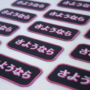May include: Multiple black fabric patches with pink neon-style lettering that reads "よろしく" (pronounced "yoroshiku") in Japanese. The patches are rectangular and have a pink border.