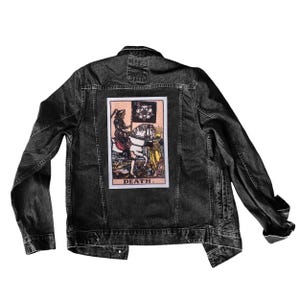 May include: Black denim jacket with a tarot card patch on the back. The patch shows a figure on horseback, with the word "DEATH" beneath. The jacket has a button-up front and two chest pockets. The jacket is made of denim.