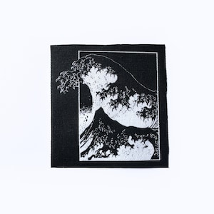 May include: A square, black fabric patch featuring a white graphic of a large ocean wave with a mountain in the background. The wave design is enclosed within a white rectangular border. The patch is on a white background.